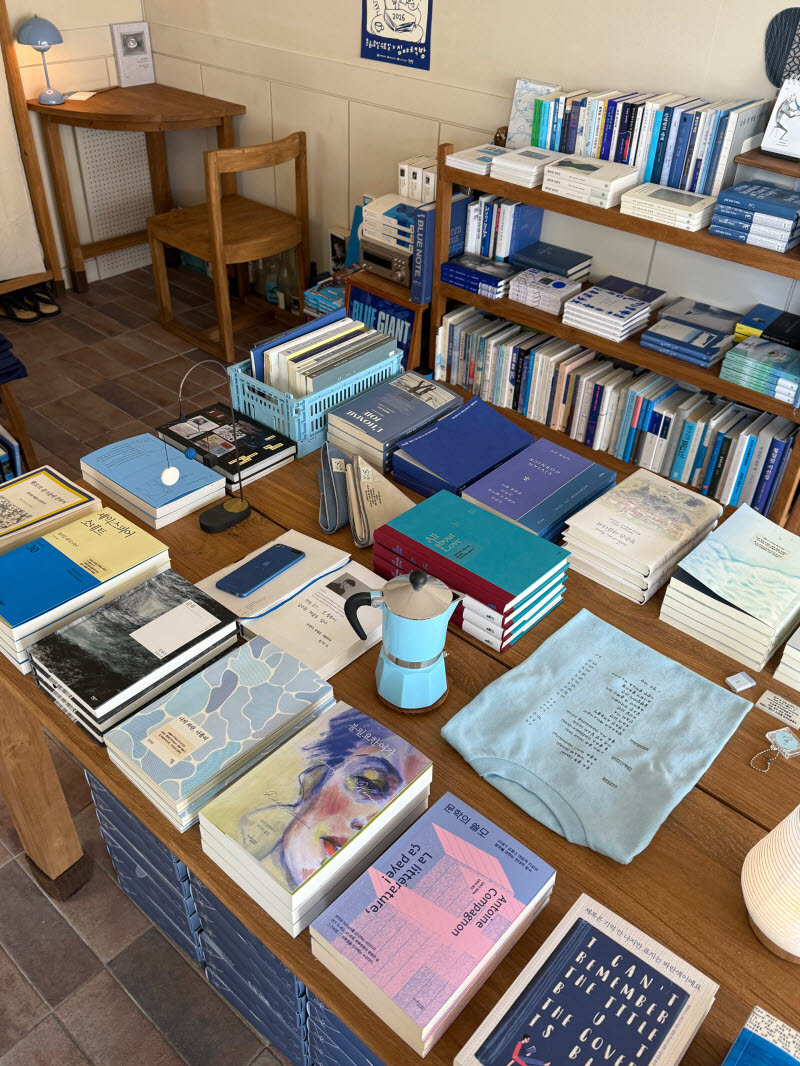 중앙 테이블 위에 한국 문학, 번역 소설, 에세이 등 다양한 장르의 책들이 펼쳐져 있다. 파란 모카포트와 하늘색 티셔츠도 책들 사이에 자연스럽게 어우러지며, 파랑으로 통일된 큐레이션의 일관성이 돋보인다.(사진=신문경 기자)